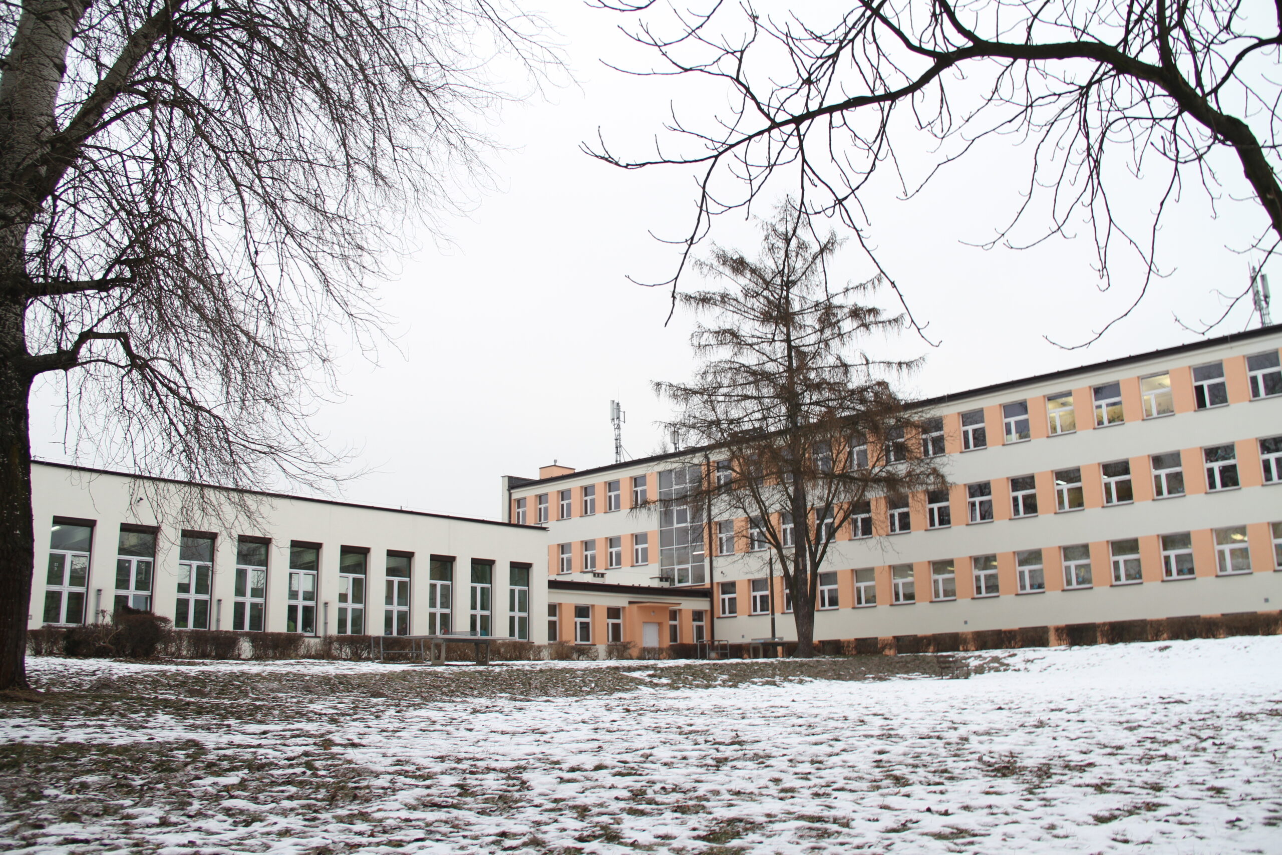 Zdjęcie przedstawia fotografię szkoły od strony wewnętrznej podczas zimy. Po lewej znajduje się sala gimnastyczna, a po prawej budynek szkoły.